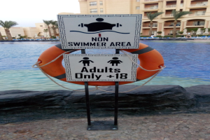 Ein Schwimmbad mit einem "Nur für Erwachsene - Nichtschwimmerbereich"-Schild im Vordergrund, umgeben von Bäumen, Gebäuden und einem klaren blauen Himmel, mit einem Rettungsring in der Mitte des Pools.