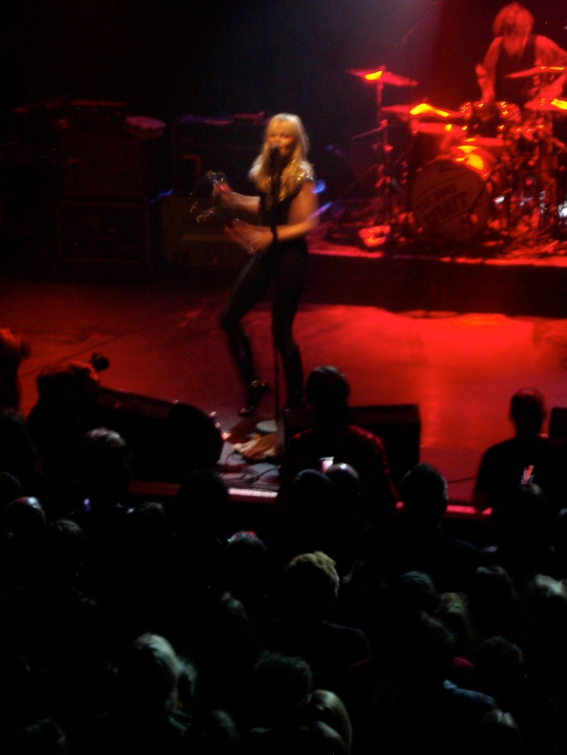 Eine Frau singt auf der Bühne in ein Mikrofon, während ein Mann daneben Schlagzeug spielt und ein Publikum im Hintergrund zu sehen ist.