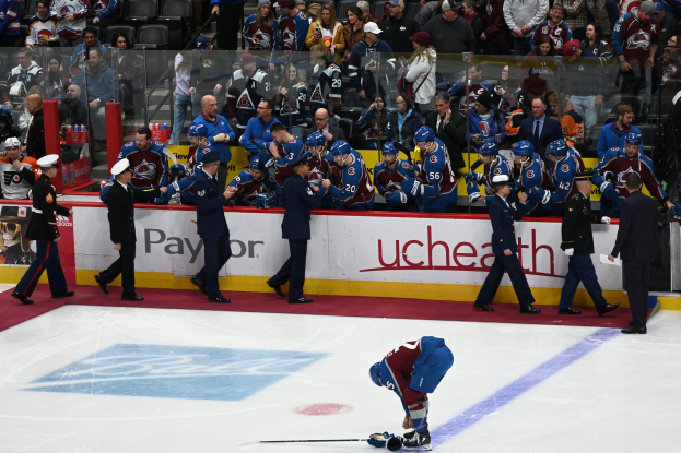 Eishockeyspieler auf dem Eis umgeben von Menschen in Mützen und mit Stöcken in der Hand, mit Zuschauern im Hintergrund und Tafeln mit der Aufschrift "Colorado Avalanche vs San Jose Sharks - 3/24/15 NHL Pick, Odds, and Prediction".