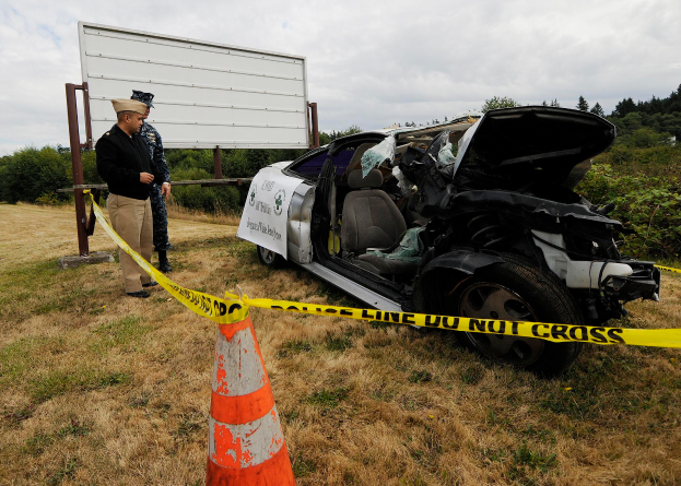 Ein Polizist in Uniform steht neben einem schwer beschädigten Auto am Straßenrand, mit einem Verkehrskegel und einem Band an einem Pfahl in der Nähe, Bäume und ein bewölkter Himmel im Hintergrund.