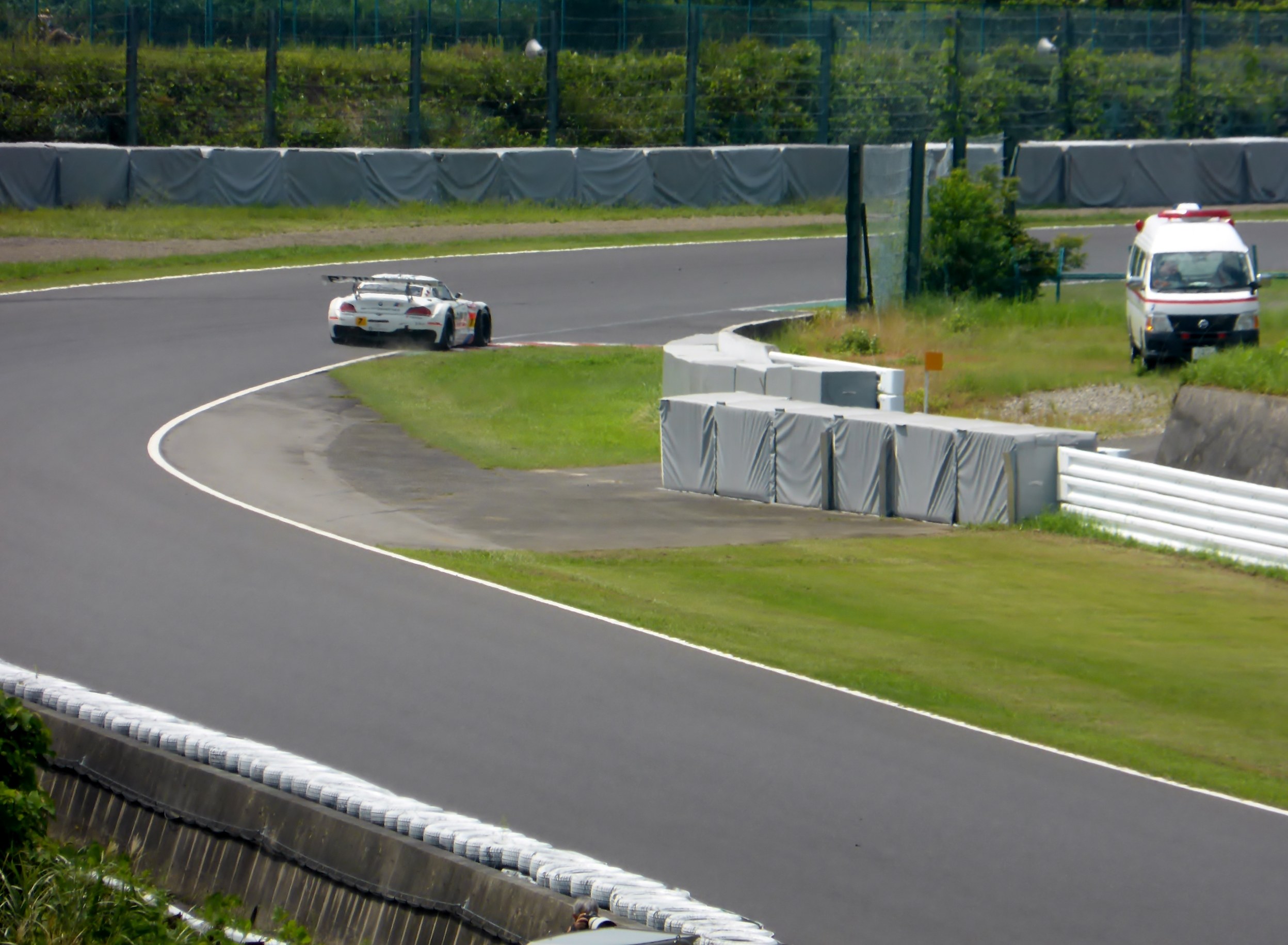 Ein Rennwagen fährt auf einer Rennstrecke mit einem Polizeiwagen im Hintergrund, umgeben von Gras, Pflanzen, Bäumen und Absperrungen und einem Fahrzeug auf der rechten Seite des Bildes.