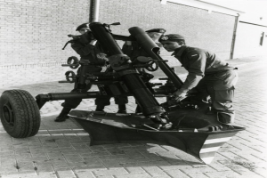 Eine Gruppe uniformierter Männer in Mützen steht neben einem schwarz-weißen Artilleriegeschütz auf einem Fahrzeug, mit einer Wand und einem Rohr im Hintergrund.