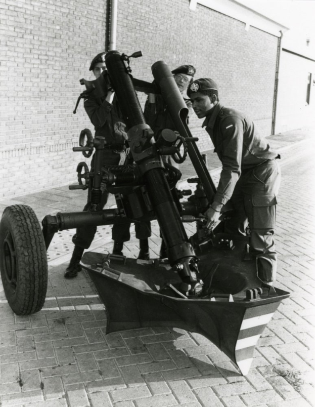 Eine Gruppe uniformierter Männer in Mützen steht neben einem schwarz-weißen Artilleriegeschütz auf einem Fahrzeug, mit einer Wand und einem Rohr im Hintergrund.