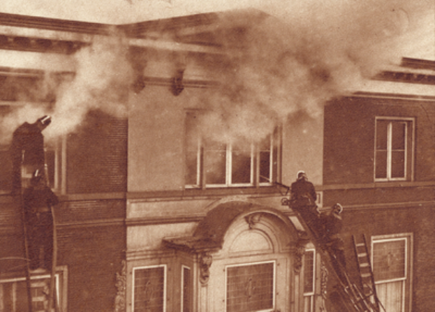Zwei Männer auf Leitern arbeiten an einem Gebäude, aus dessen Fenstern Rauch quillt, dargestellt in Schwarz-Weiß.