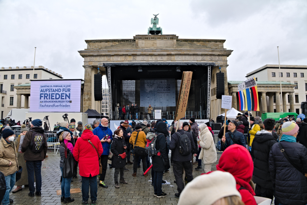 Eine Menschenmenge steht vor einem Gebäude mit einer Bühne, Rednern und einem Bildschirm, hält Fahnen und Schilder, was auf eine Demonstration in Berlin hindeutet.