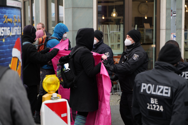 Eine Gruppe von Menschen steht vor einem Gebäude mit Glaswänden und Deckenbeleuchtung, einige tragen Mützen und tragen Taschen oder Kleidung, während ein Polizist in Berlin, Deutschland, eine Frau festnimmt; ein Pfahl mit Text und ein Wasserzeichen sind sichtbar.
