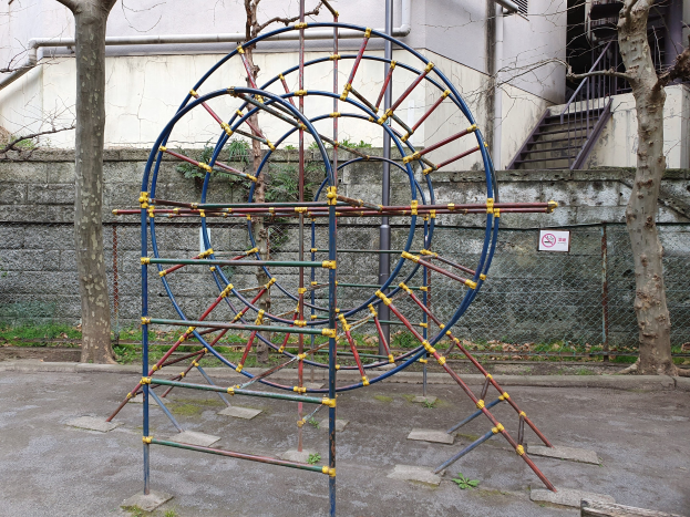 Spielplatz mit Metall-Klettergerüst, umgeben von Bäumen und einer Mauer mit Maschendrahtzaun, mit Treppen, die zu einem Gebäude mit Fenstern und Rohren führen, einem Schild und Grasboden.