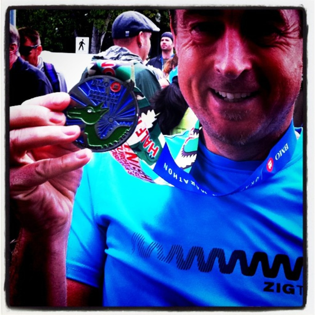 Ein Mann lächelt und hält eine Medaille in der Hand, umgeben von anderen Menschen und einer Tafel mit Blättern.