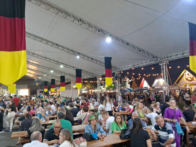 Große Gruppe von Menschen an langen Tischen unter einem Outdoor-Tent auf dem Oktoberfest in München, Deutschland, mit weiteren Gästen in der Nähe, Taschen und Gegenständen auf den Tischen, festlichem Hintergrund von Zelten, Lichtern und Fahnen.