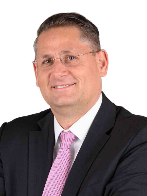 Ein Mann in Anzug und Krawatte, der lächelt und eine Brille trägt, vor einem weißen Hintergrund.