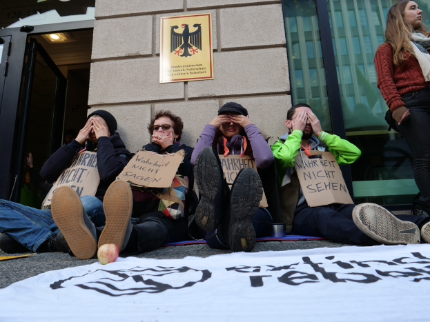 Eine Gruppe von Menschen sitzt vor einem Gebäude auf dem Boden und hält Schilder, mit einer Fahne und einem Apfel auf der Straße und einer Frau, die rechts steht und besorgt aussieht.
