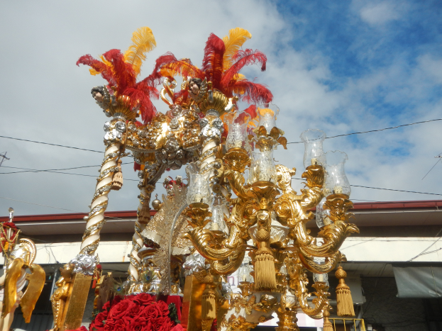 Ein großer goldener und roter Festwagen mit Blumen und anderen Schmuckelementen bei einem Faschingsumzug, mit einem Gebäude, Strommasten mit Drähten und einem bewölkten Himmel im Hintergrund.