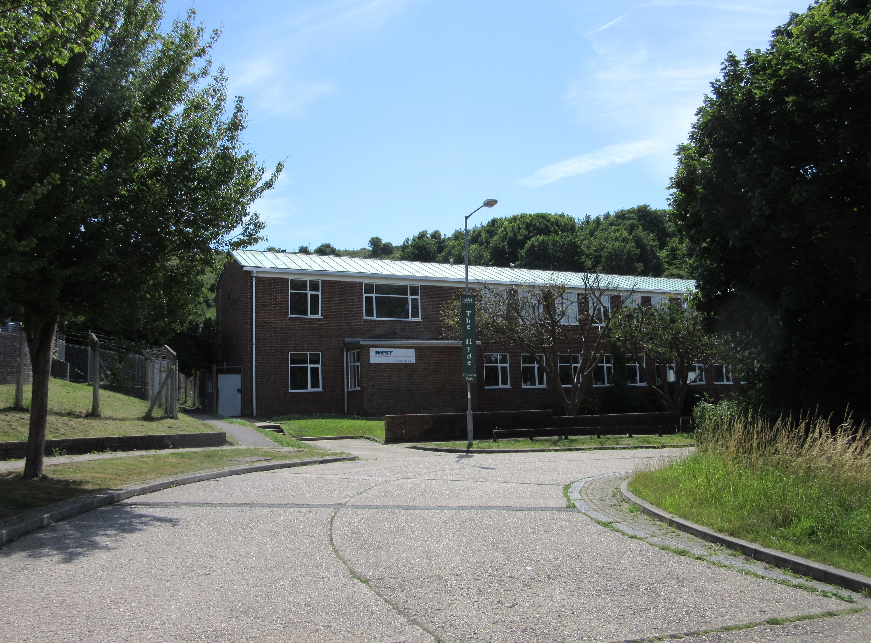 Ein Schulgebäude mit Fenstern, einer Straßenlaterne, einem Schild, einer Baumgruppe, Gras, einem Zaun und einem bewölkten Himmel, das in der Mitte eines Parks umgeben von saftigem Gras liegt.