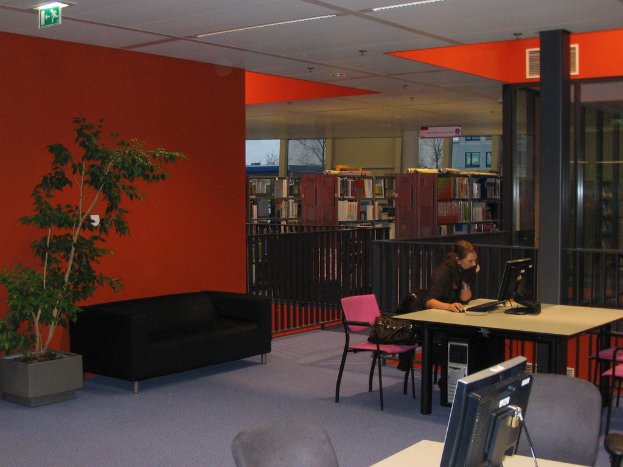 Eine Frau in einem schwarzen T-Shirt bedient ein Computersystem auf einem Tisch, mit einer schwarzen Couch dahinter, einer roten Wand mit einer Pflanze und Bücherregalen im Hintergrund, alles unter einer Decke mit Fenstern, die ein Gebäude draußen zeigen.