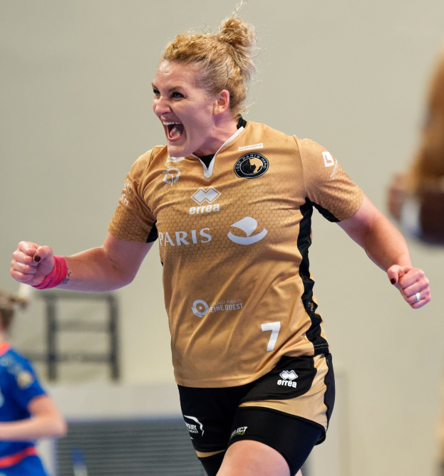 Eine Frau in braunem T-Shirt und schwarzen Shorts rennt und feiert auf einem Futsalplatz, mit zwei anderen Frauen im Hintergrund.