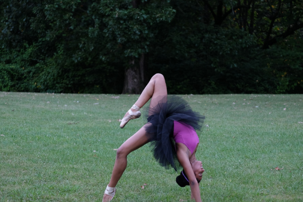 Eine Frau in einem rosafarbenen und schwarzen Kleid führt Turnübungen auf Gras mit ein paar Bäumen im Hintergrund aus.