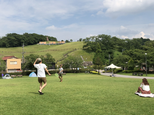 Eine Gruppe von Menschen, die Badminton in einem Park spielt, mit Zelten, Straßenlaternen, Strommasten, Stromkabeln, Gebäuden, Bäumen, Hügeln und einem bewölkten Himmel im Hintergrund.