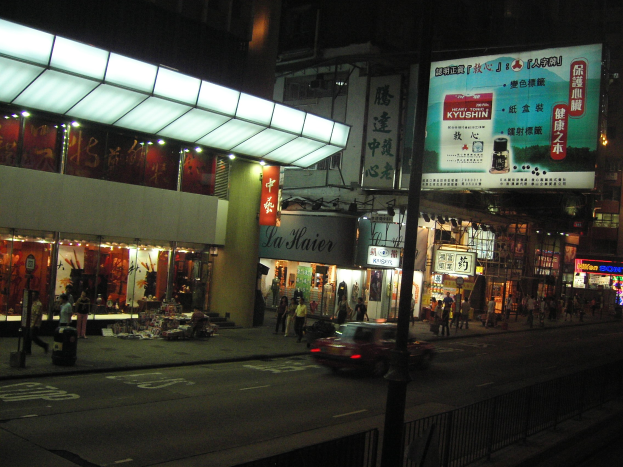 Eine belebte Stadtstraße bei Nacht mit einem Auto, das die Straße entlangfährt, umgeben von Gebäuden, Geschäften, Namensschildern, Strommasten, Kabeln, Geländern und Fußgängern auf dem Gehweg.
