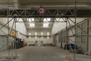 Großes Lagerinnere mit ausgedehnten Gerüsten, Verkehrskegeln, Metallstangen, einem Schild, Maschinen, einer Tür, Fenstern, Wänden, Deckenleuchten und Dach.