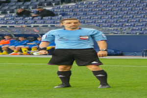 Ein Mann in einem blauen Hemd und schwarzen Shorts steht auf einem Fußballfeld, umgeben von sitzenden und stehenden Menschen, die Fußballschuhe tragen.