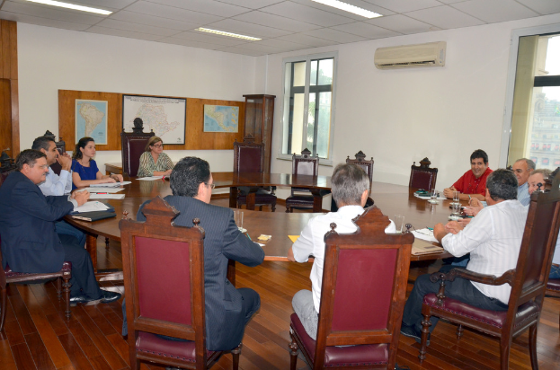 Eine Gruppe von Menschen, die an einem Tisch mit Papieren und Gläsern sitzen, ein Klimagerät, ein Anschlagbrett und Karten an der Wand im Hintergrund.