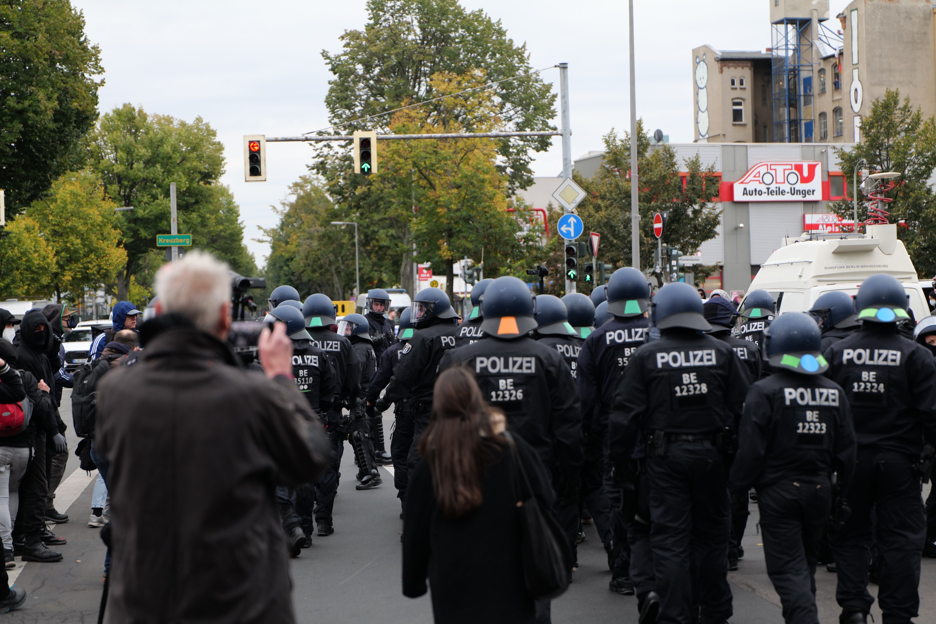 Eine Gruppe von Menschen geht eine Straße entlang, mit Polizeibeamten, einige tragen Helme und tragen Taschen oder Kameras, mit Fahrzeugen, Verkehrszeichen, Schildern, Bäumen, Gebäuden und einem klaren blauen Himmel im Hintergrund.