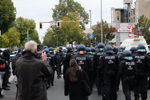 Eine Gruppe von Menschen geht eine Straße entlang, mit Polizeibeamten, einige tragen Helme und tragen Taschen oder Kameras, mit Fahrzeugen, Verkehrszeichen, Schildern, Bäumen, Gebäuden und einem klaren blauen Himmel im Hintergrund.