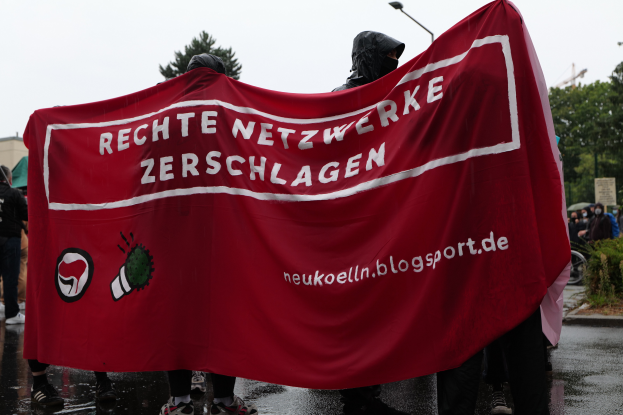 Eine Gruppe von Menschen steht auf einer Straße und hält ein rotes Banner mit der Aufschrift "Rechte Netzwerke Zerschlagen" in den Händen, umgeben von Bäumen, Gebäuden, Laternenmasten und einem klaren blauen Himmel, eine Person fährt rechts auf einem Fahrrad und eine weitere Person hält links einen Schirm.