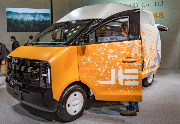 Orange und weißer Elektro-Transporter auf einer Ausstellung präsentiert, umgeben von Menschen in bunten Kleidern, mit einer Wandmontage und Text im Hintergrund.