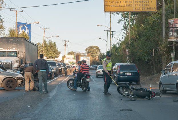 Eine Gruppe von Menschen umringt ein verunglücktes Motorrad am Straßenrand mit vorbeifahrenden Fahrzeugen, Bäumen, Strommasten und einem klaren Himmel im Hintergrund.