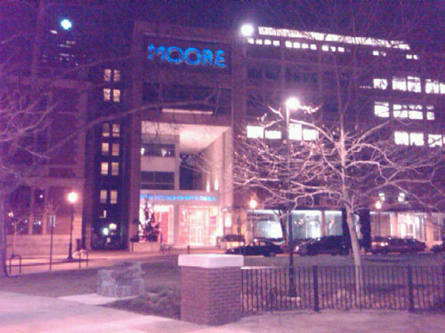 Ein Krankenhausgebäude mit mehreren Fahrzeugen, Bäumen und einem Zaun davor.