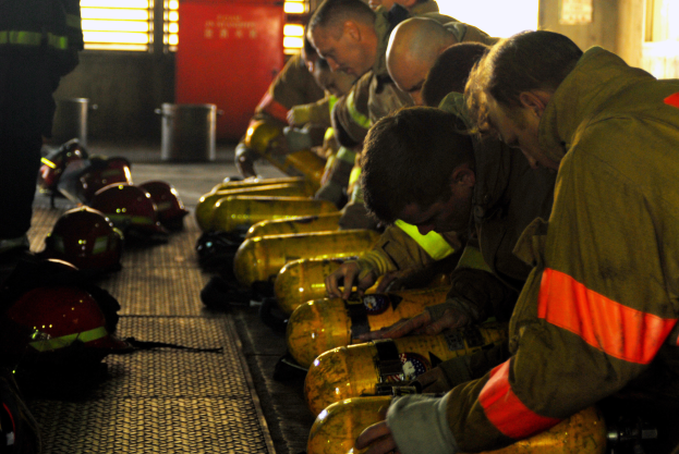 Eine Gruppe von Feuerwehrleuten bei der Arbeit an einem Feuerwehrauto mit verstreuten Helmen und Zylindern auf dem Boden, mit einer Wand mit Fenstern und einer Tür sowie einem Müllcontainer im Hintergrund.