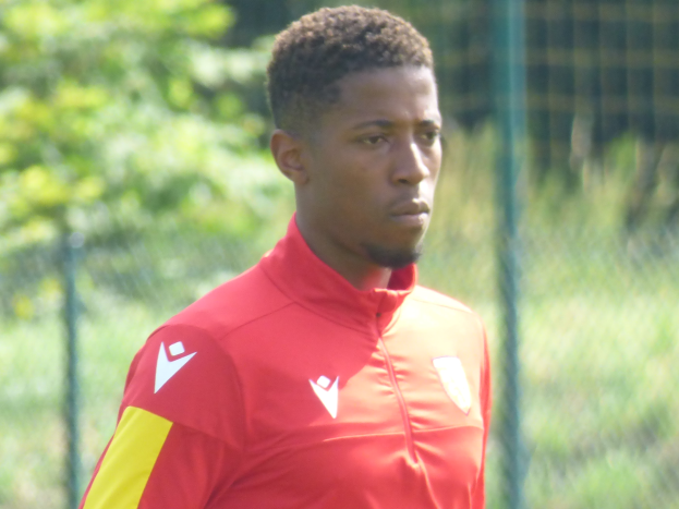 Ein junger Mann in einem roten und gelben Trikot steht auf einem Fußballfeld umgeben von einem Zaun und Bäumen und sieht entschlossen aus während eines Trainings.