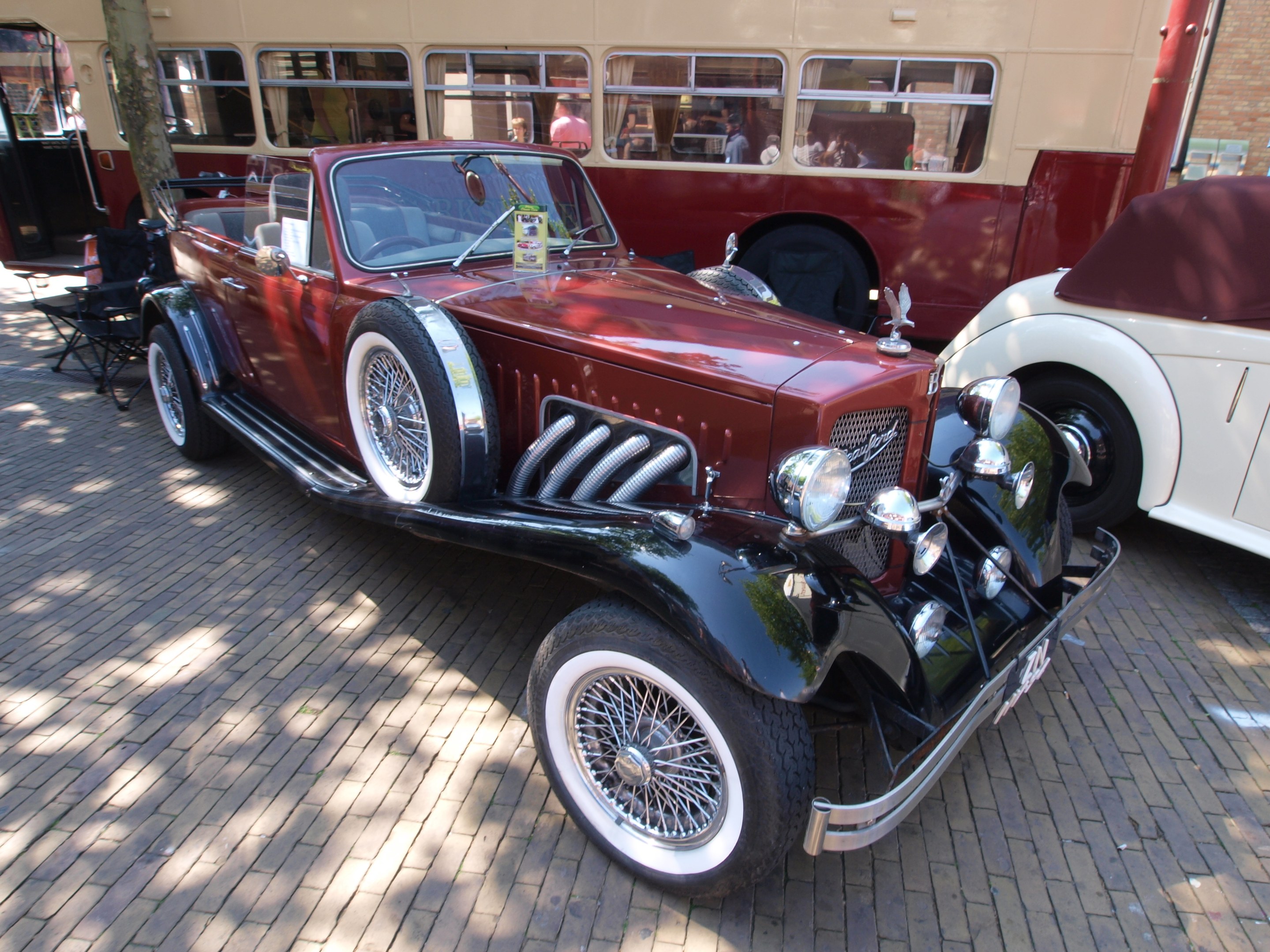 Vintage-Auto neben einem Doppeldeckerbus auf einem Kopfsteinpflasterweg geparkt, mit Stühlen und einem Baum im Vordergrund, einem Gebäude mit Fenstern im Hintergrund und Menschen im Bus sichtbar.