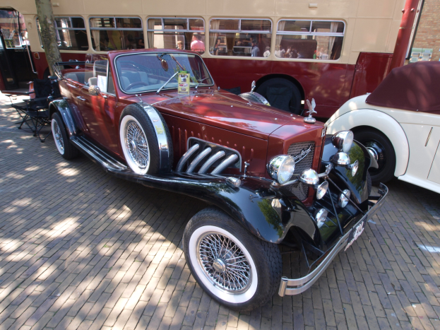Vintage-Auto neben einem Doppeldeckerbus auf einem Kopfsteinpflasterweg geparkt, mit Stühlen und einem Baum im Vordergrund, einem Gebäude mit Fenstern im Hintergrund und Menschen im Bus sichtbar.