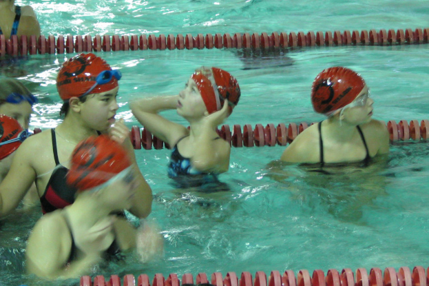 Kinder in einem Schwimmbad, alle mit Badekappen und Schwimmbrillen, mit Wasser im Hintergrund.