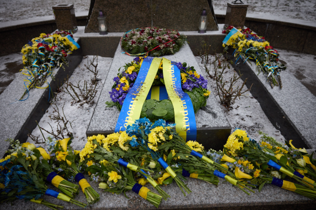 Gedenkstätte in Kiew, Ukraine, mit einem Blumenkranz auf einem Grab, das von einer blauen Schleife markiert ist, umgeben von zusätzlichen Blumensträußen, vor einem verschneiten Hintergrund.