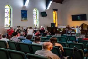 Eine Gruppe von Menschen sitzt auf Stühlen in einer Kirche, ein Mann steht vorne mit einem Mikrofon in der Hand, umgeben von Musikinstrumenten, einem Tisch mit einer Vase, einem Lautsprecher, einer Wandbanner mit Text, Fenstern und Deckenleuchten.