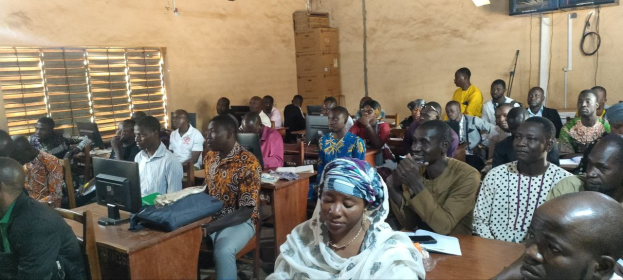 Eine Gruppe von Menschen, die an Tischen in einem Klassenzimmer sitzen, mit Monitoren, Taschen, Büchern und Papieren, an einer Techniktrainingssitzung teilnehmen; ein Fenster auf der linken Seite, Kartons und ein Fernseher an der Wand im Hintergrund.