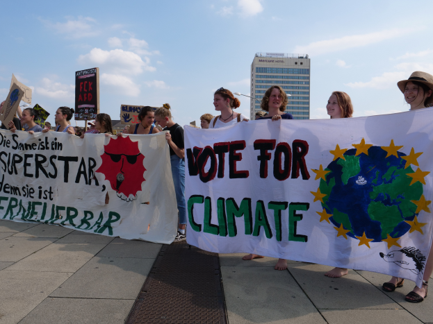 Eine Gruppe von Menschen mit einer 'Wähle für das Klima'-Plakette und verschiedenen Schildern, die auf dem Boden stehen, mit Gebäuden, Straßenlaternen, Straßenlaternen und einem bewölkten Himmel im Hintergrund.