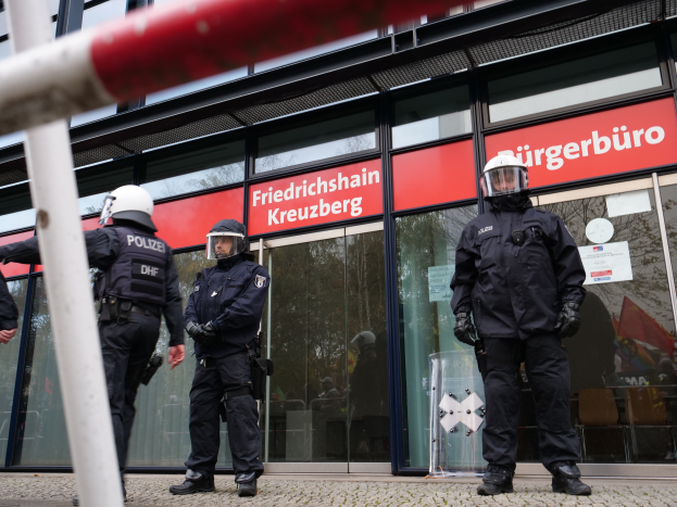 Eine Gruppe von Polizisten in Helmen und Handschuhen steht vor einem Glaswandgebäude mit aufgeklebten Papieren, einem Pfahl links und Schildern mit der Aufschrift "Friedrichshain Kreuzberg" im Hintergrund.