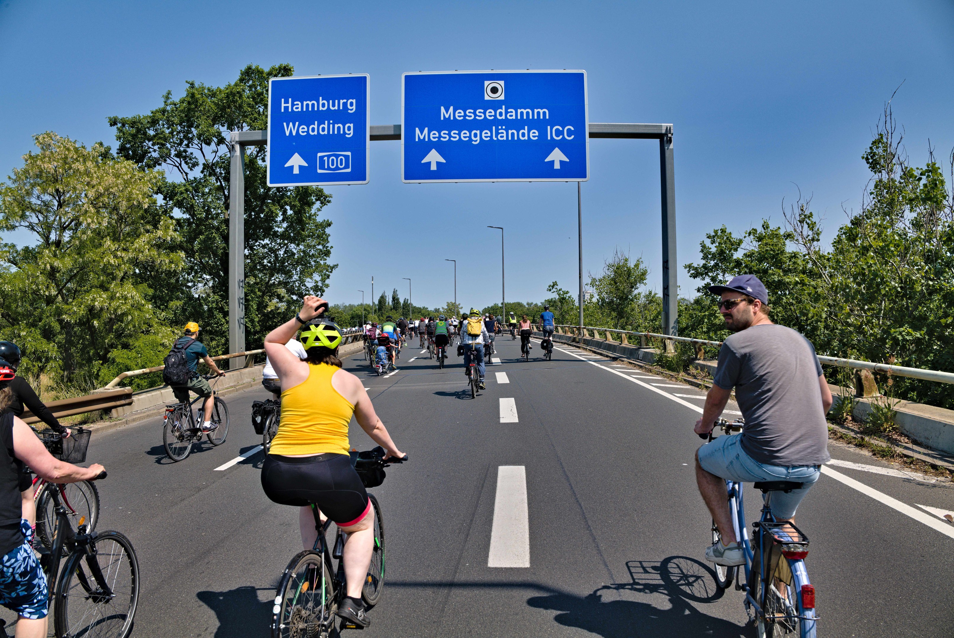 Gruppe von Radfahrern mit Helmen auf einer Straße mit Bäumen auf einer Seite und einem Geländer auf der anderen, Laternenmasten und einem klaren blauen Himmel im Hintergrund, mit einer Tafel, die eine Radtour in Hamburg anzeigt.