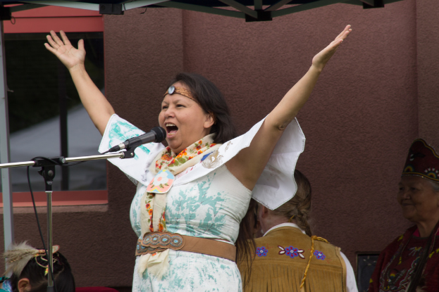 Eine Frau in einem weißen Kleid steht auf einer Bühne, schreit in ein Mikrofon, mit einer Wand und einem Eisenrahmen im Hintergrund.