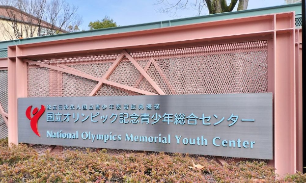 Außenansicht des Nationalen Olympischen Memorial Youth Center mit einem Namen auf der Wand, Pflanzen am Boden, Bäumen und Gebäuden im Hintergrund und einem klaren blauen Himmel darüber.