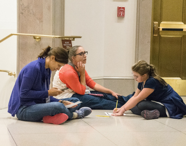 Drei Frauen sitzen auf dem Boden vor einer Tür, umgeben von Büchern und Schreibmaterialien, in Diskussion vertieft.