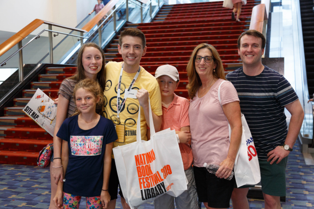 Eine Gruppe von Menschen, die zusammenstehen, lächeln und Taschen halten, vor einer Rolltreppe auf dem Nationalen Buchfest 2019, mit Treppen, Geländern und einer Wand im Hintergrund.