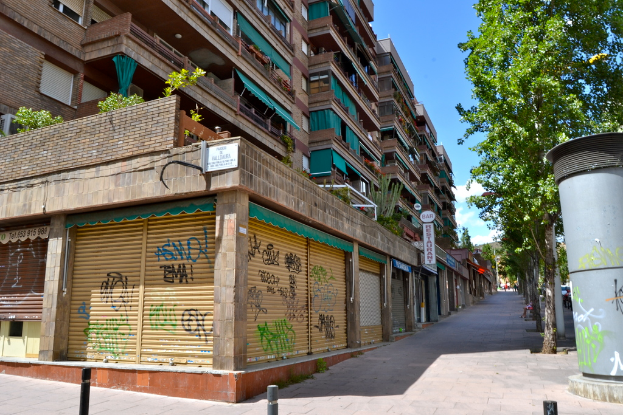 Eine Szene mit mehreren Gebäuden, Fenstern, grünen Kleidungsstücken, Geschäften, Schildern und umliegenden Bäumen und Pflanzen.