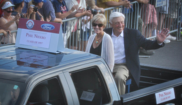 Ein Mann und eine Frau sitzen im hinteren Teil eines Fahrzeugs auf einer Straße, winken und lächeln, während eine Menge von Menschen im Hintergrund Fotos macht, wobei eine Absperrung auf der Seite der Straße sichtbar ist.