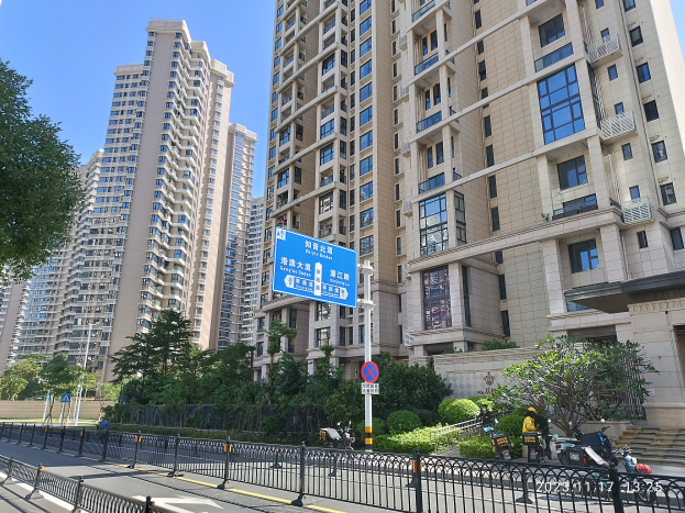 Eine Stadtstraße in Hong Kong mit hohen Wohngebäuden, umgeben von Grün und einem Zaun, mit Schildern, Pfählen, Fahrzeugen und einem klaren blauen Himmel.