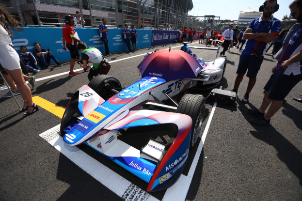 Formula E Auto mit weißer, blauer, roter und schwarzer Lackierung auf einer Rennstrecke geparkt, umgeben von Menschen, mit einem Schirm im Vordergrund und einem klaren blauen Himmel im Hintergrund.